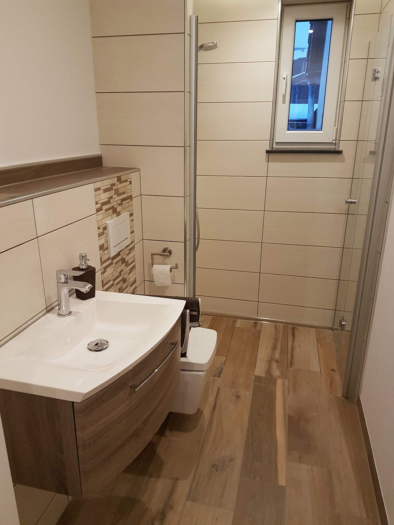 Modern bathroom: wooden vanity with white ceramic basin, glass shower, toilet, window, and beige tiles.