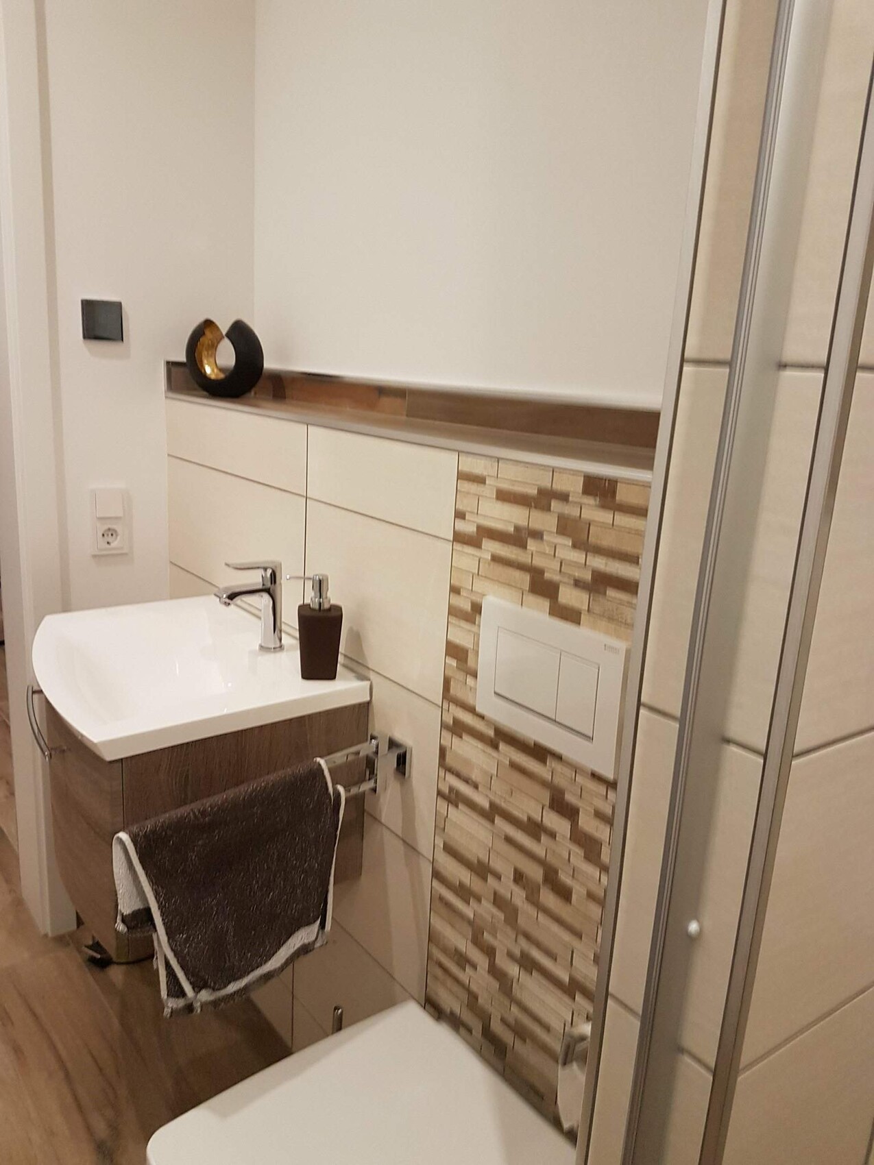 Modern bathroom with sink, wooden cabinet, mosaic wall, towel, toilet cistern, and shower.