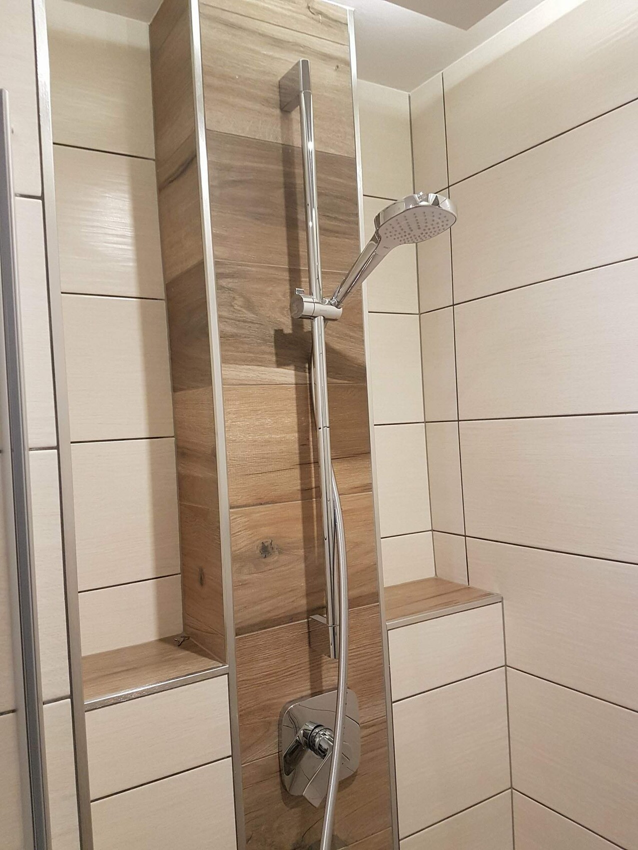 Modern shower with handheld showerhead on stainless steel rail in front of wood-effect wall panel; built-in bench.
