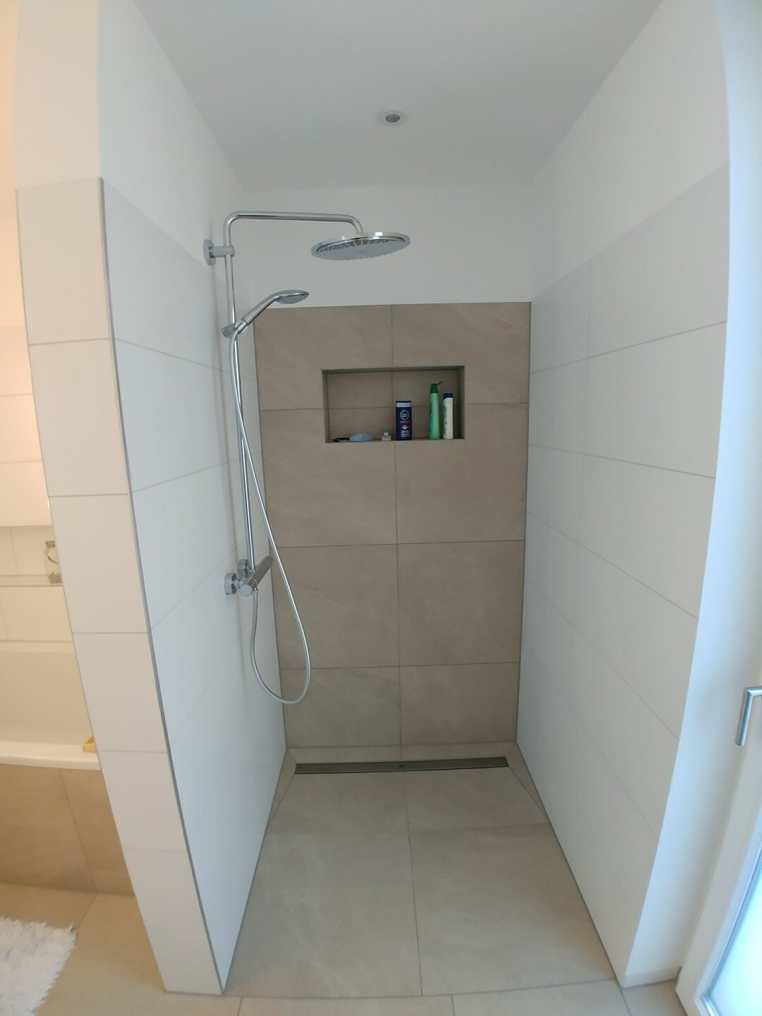 Open shower area with ceiling showerhead and handheld shower, beige tiles, niche with bottles.
