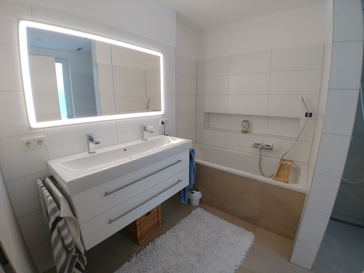 Modern bathroom scene with double sink, illuminated mirror, bathtub, and showerhead.