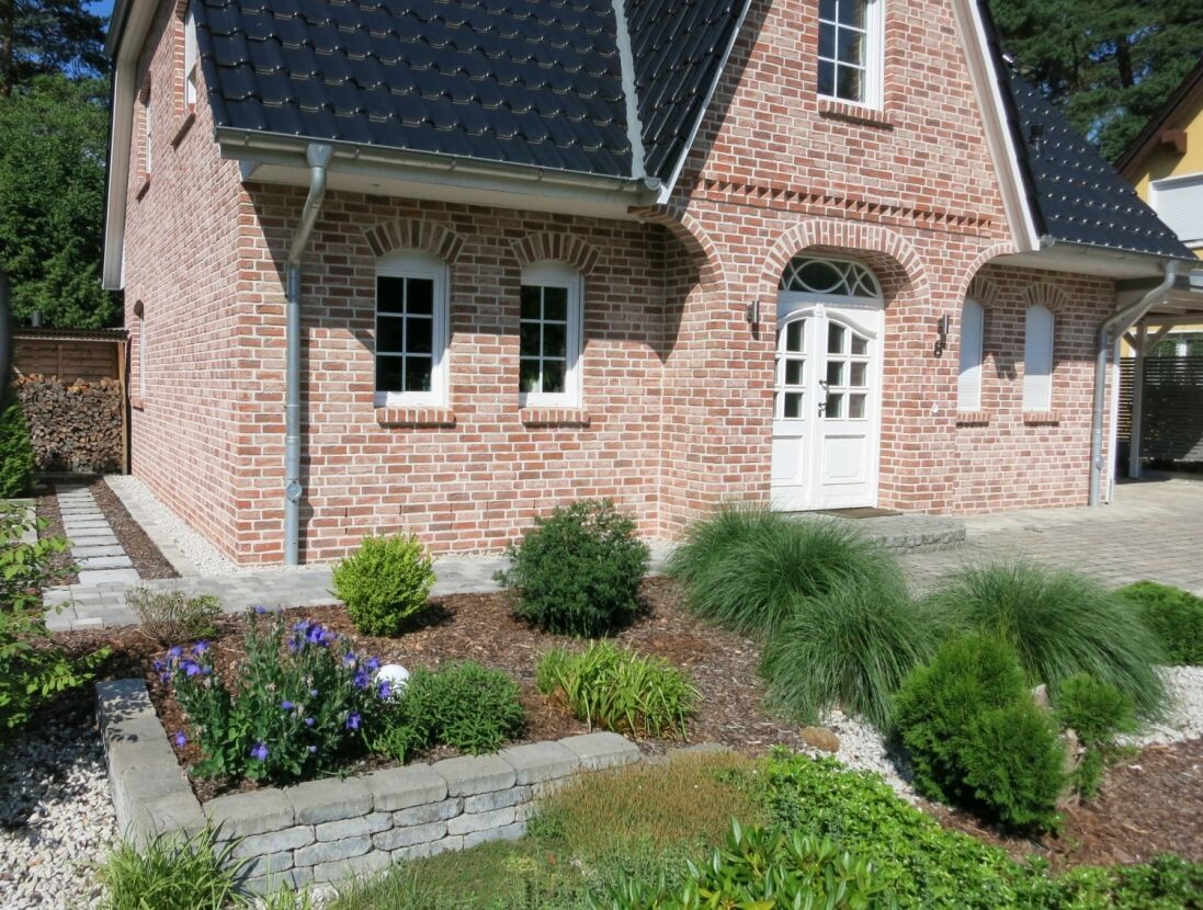 Ziegelhaus mit weißer Tür und zwei Fenstern; Vorgarten mit Grünpflanzen und Blumen.