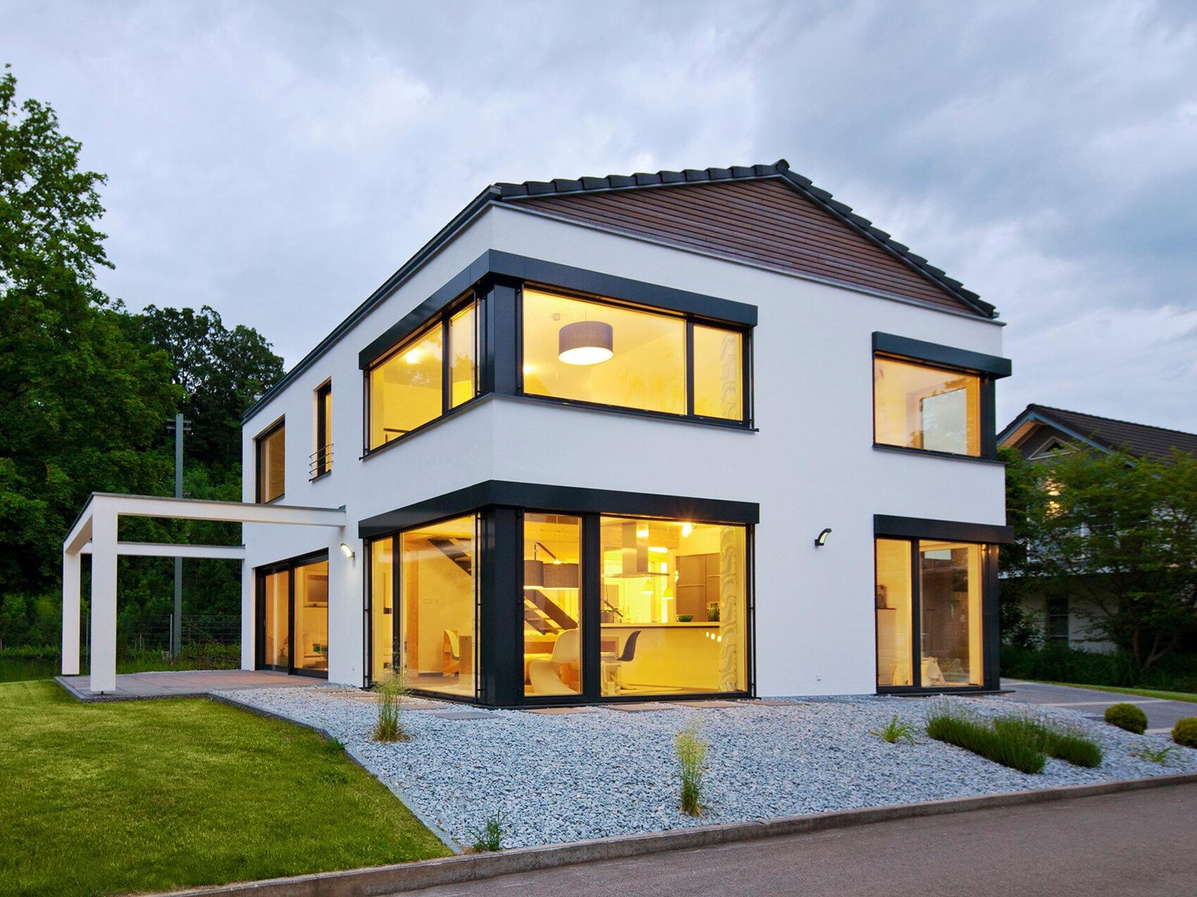 Modern white house facade with large glass fronts, illuminated interiors at dusk.