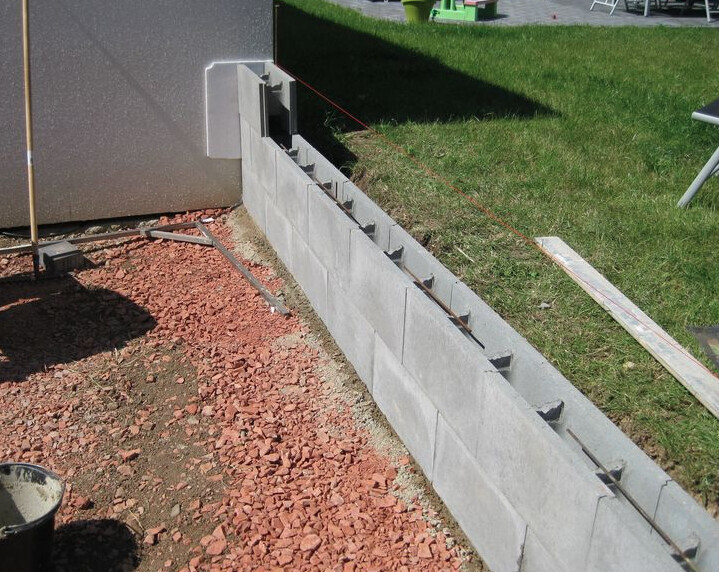 Construction site with partially built concrete block wall and reinforcement at the foundation edge.