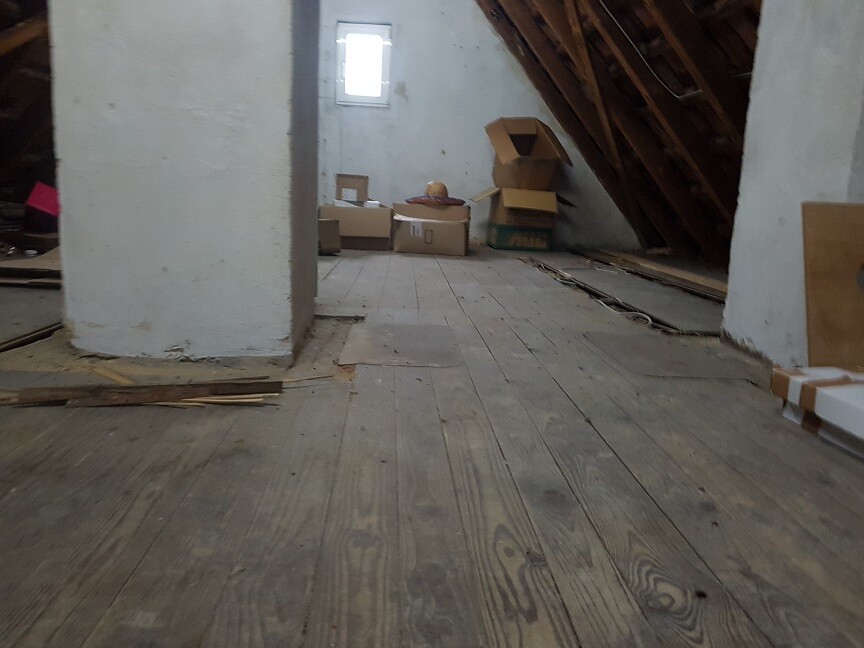 Attic with wooden floorboards, boxes, crates, and construction timber; small window in the background.