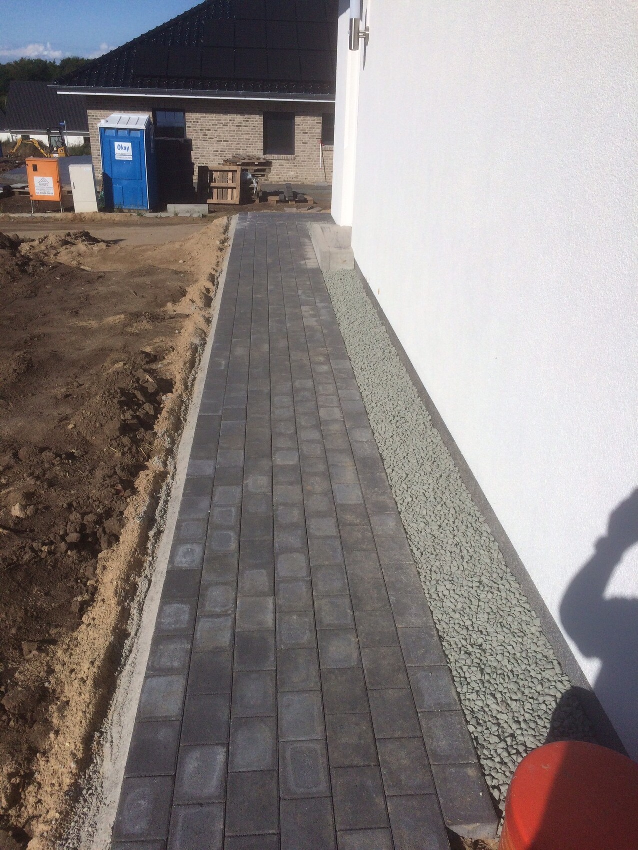 Gepflasterter Gehweg aus grauen Pflastersteinen neben einer weißen Wand; Kiesstreifen, Baustelle.