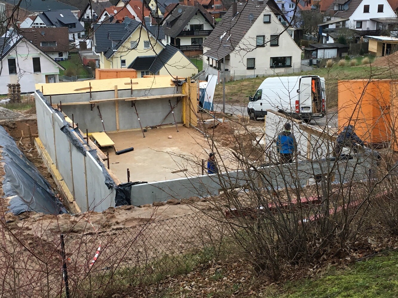 Construction site with foundations and workers, white van in front of orange container.