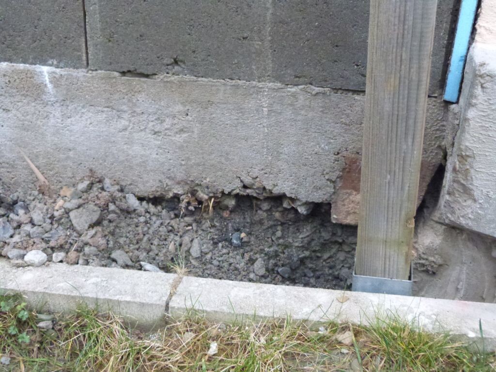 Damaged foundation area with soil, concrete wall, and wooden post