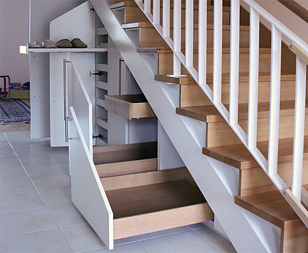 Under the staircase open storage system with drawers and doors, wooden steps, white railing.