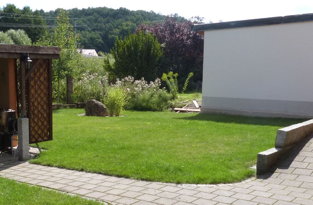 Green garden with lawn, paved path, wooden pavilion on the left, and white building on the right.