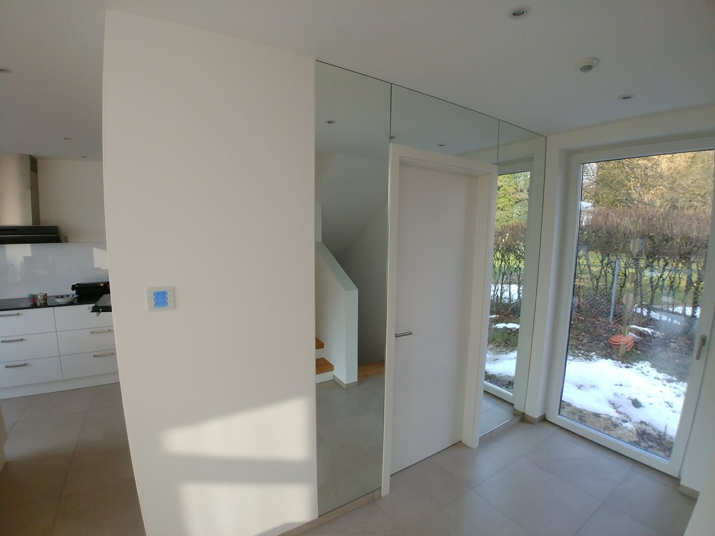 Modern white kitchen with mirrored fronts, large window overlooking snow outside.