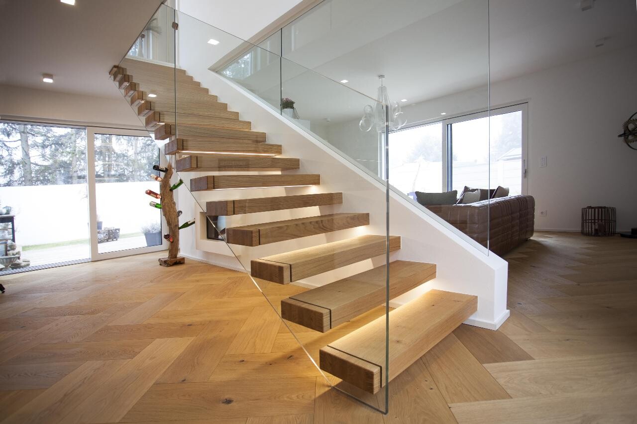 Modern, bright living room with floating wooden staircase, glass railing, and parquet floor.