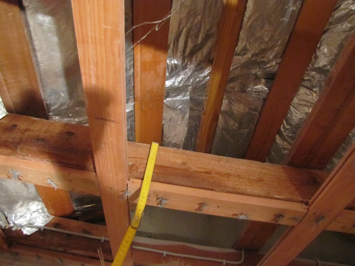 Attic with wooden beams, foil insulation and a diagonal yellow measuring tape over a beam.