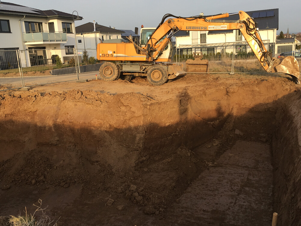 Excavator digging construction pit at site; residential houses in the background