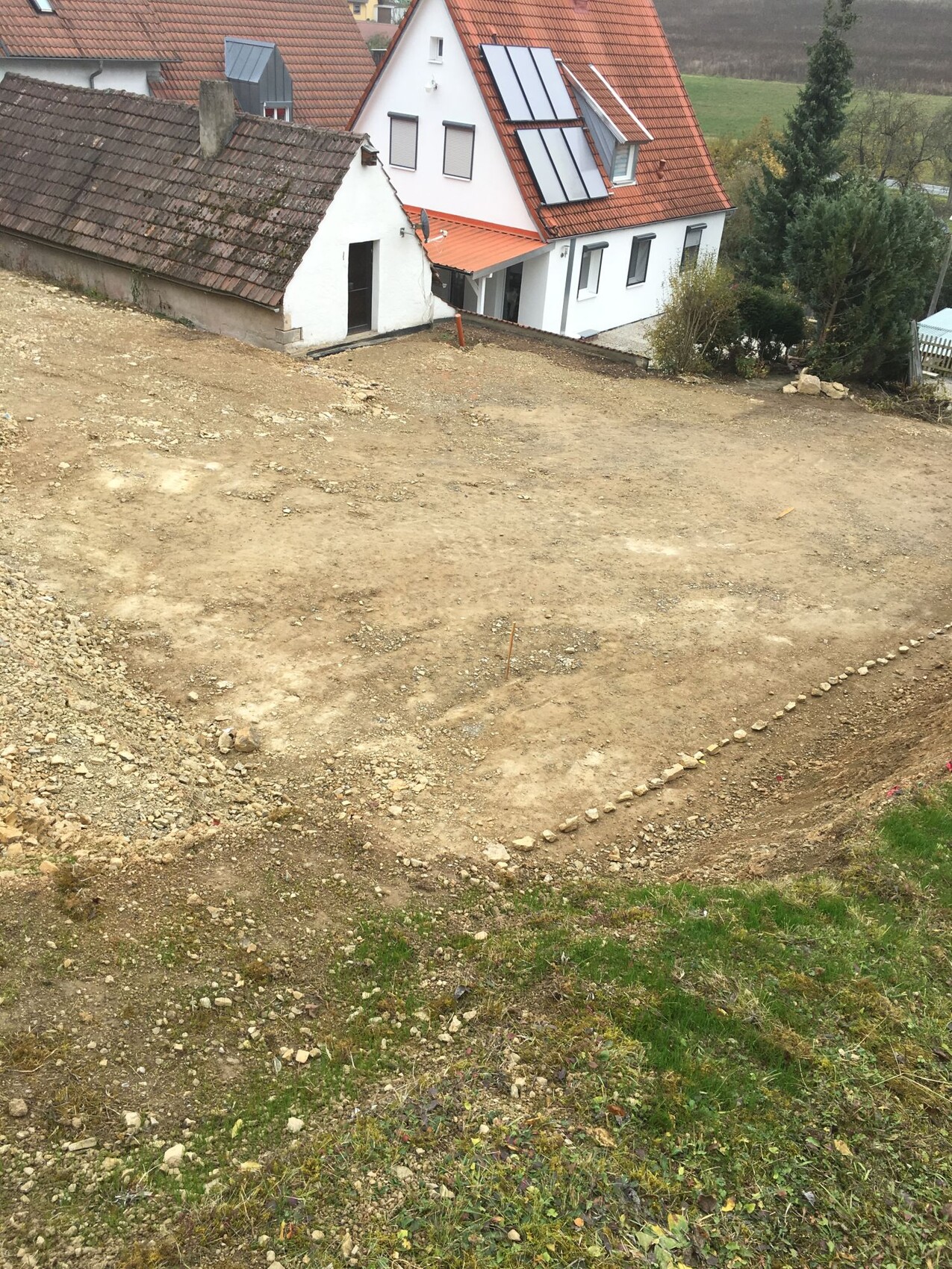 Baustelle auf Hang: Erdreich, unfertige Fundamente und weißes Haus mit rotem Dach