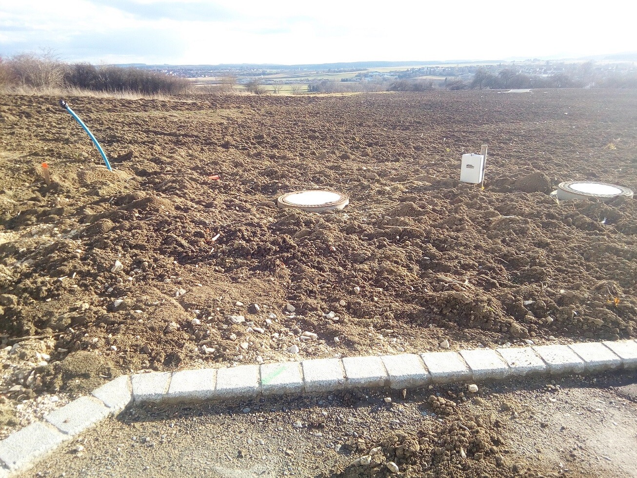 Open construction area with soil, pipes, and covers, curb at the edge.