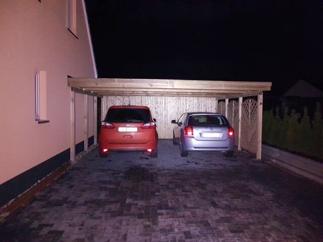 Zwei geparkte Autos unter einem Holzcarport neben einem Haus bei Nacht; roter und silberner Pkw.