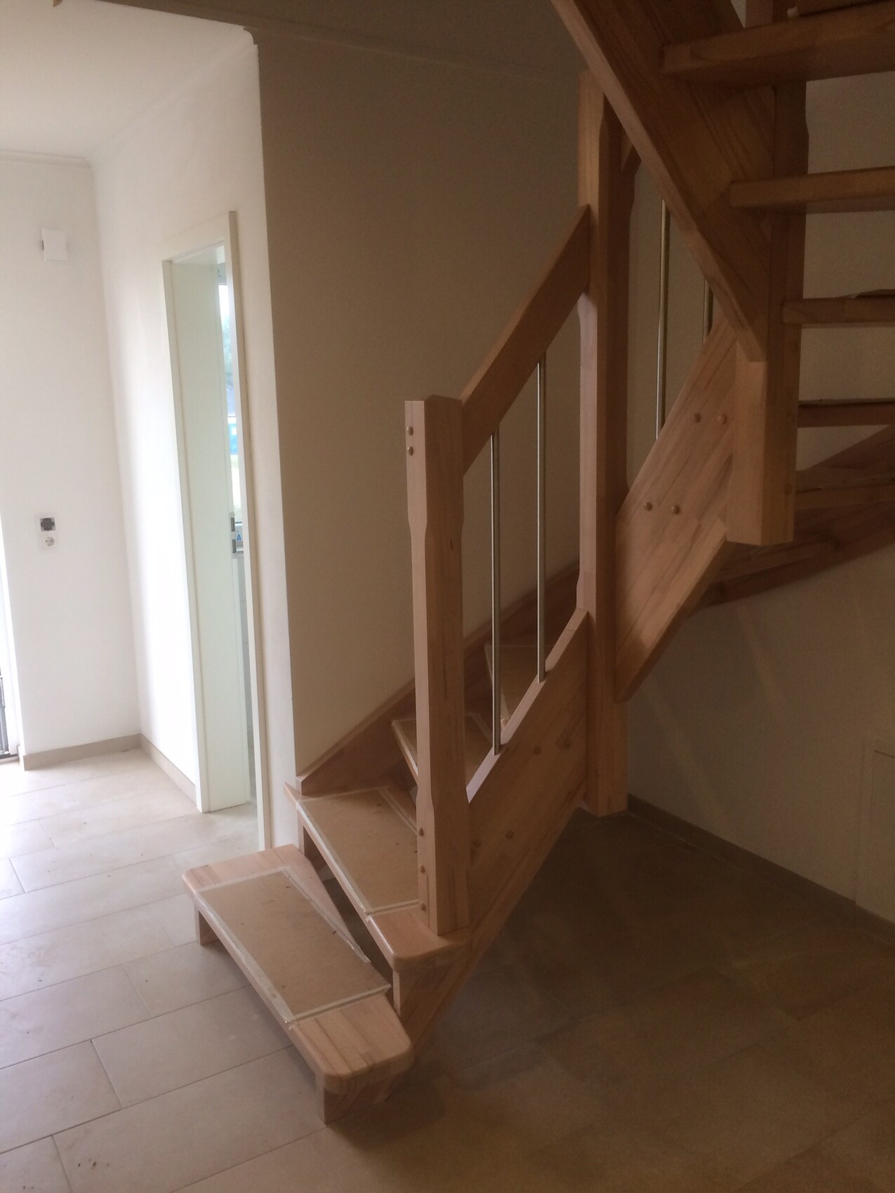 Holztreppe im Innenraum mit Geländer aus Holz und Metallstäben, Fliesenboden und hellen Wänden.