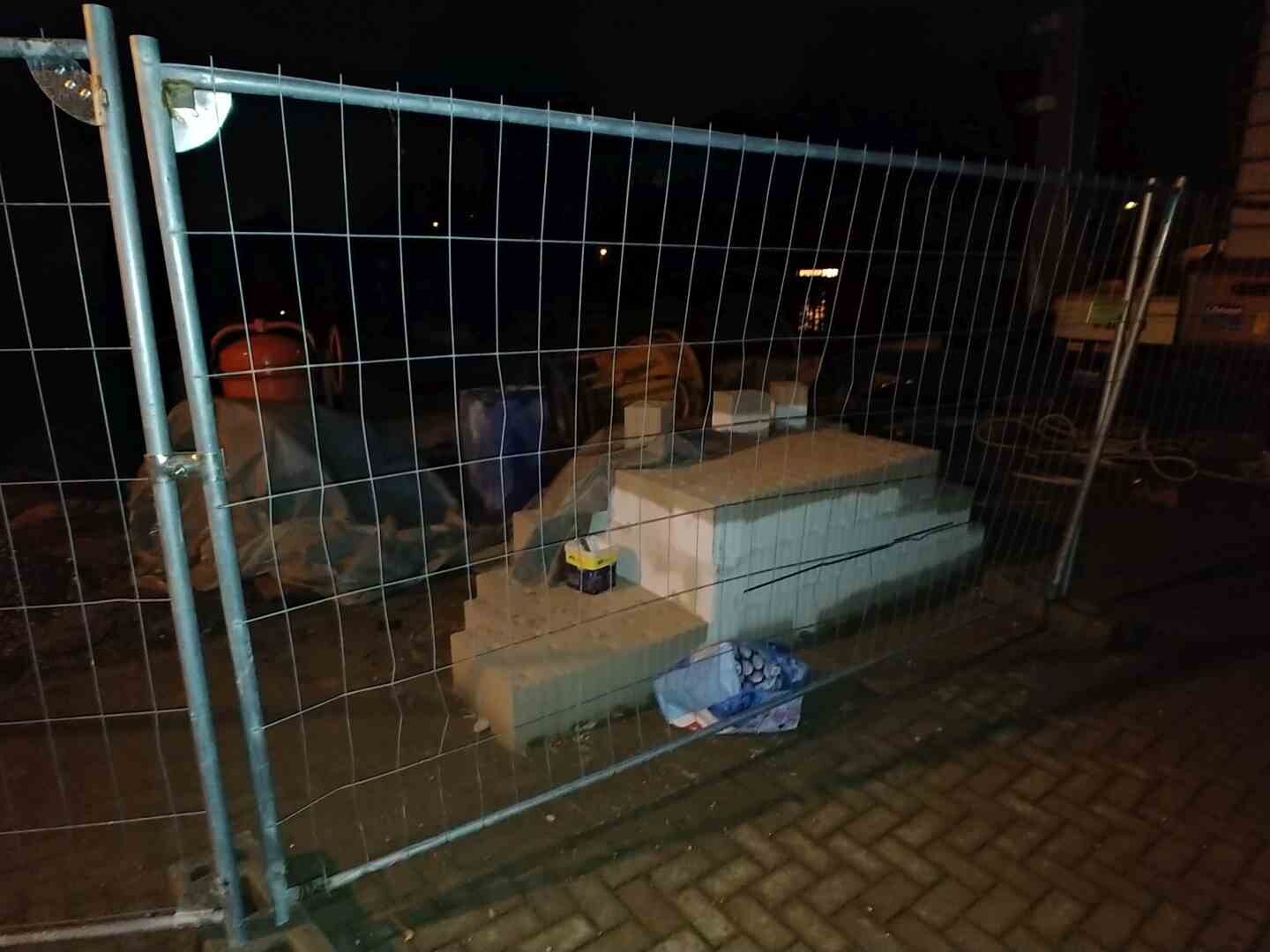 Night photo of a construction site with metal fence, building materials, and foundation stones