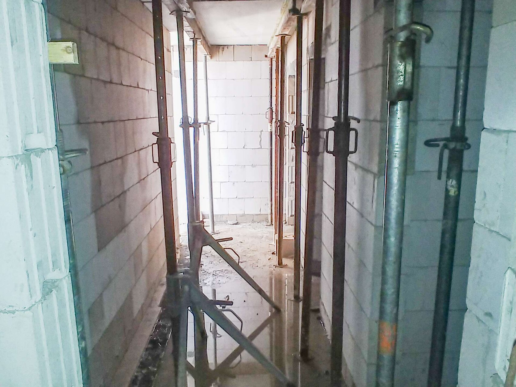 Narrow construction corridor with steel scaffolding and puddles on the floor.