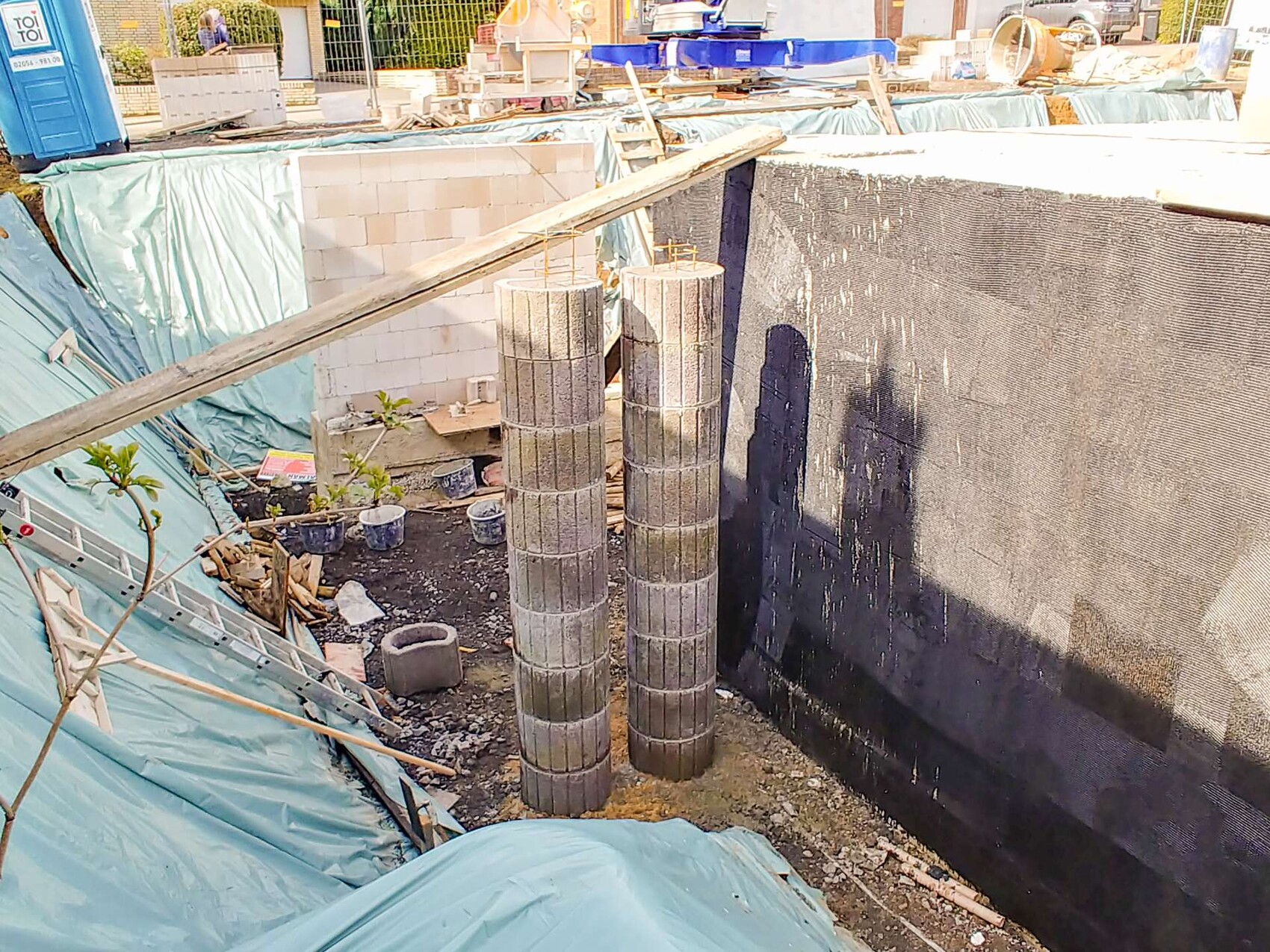 Construction site: Excavation pit with three concrete piles, tarpaulin, portable toilet and building materials.