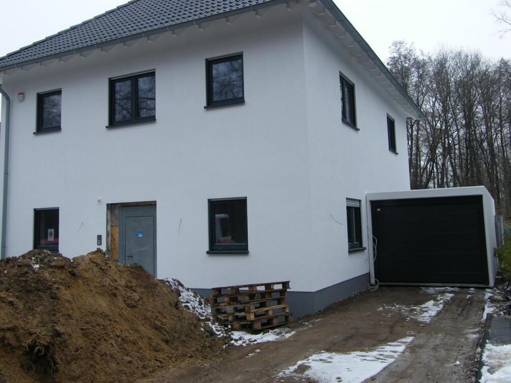 White two-story house with black garage; construction wood pallets, earth mound, and snow on the ground.