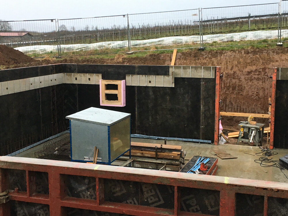 Excavation pit with black waterproofing, blue metal box, tools, and wooden battens on construction site
