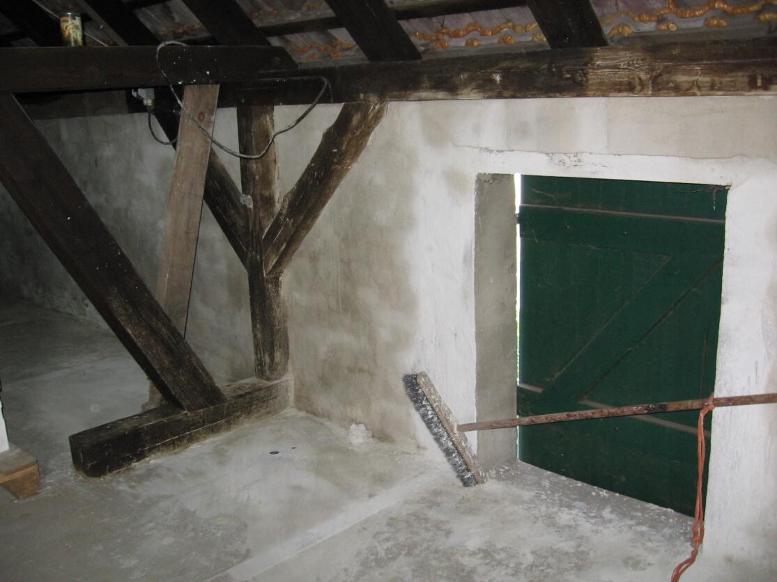 Dachboden mit dunklen Holzbalken, weißer Wand, grüner Tür und rostiger Stange mit Seil.