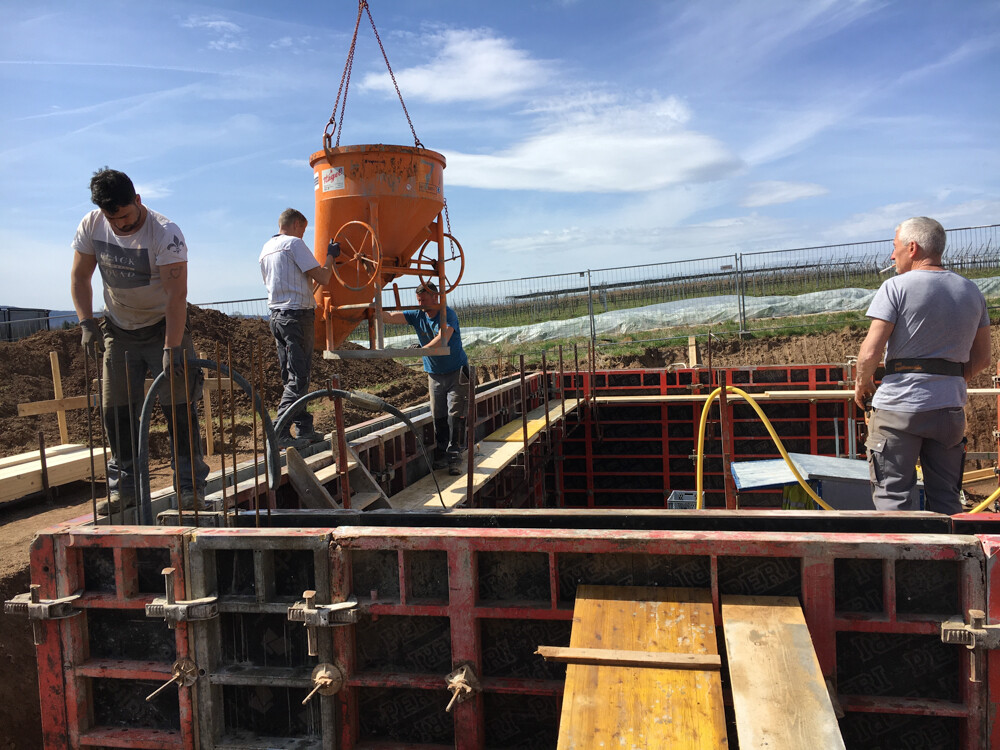Four construction workers at a building site; an orange concrete mixer being lifted over an excavation.