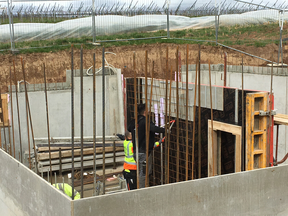Excavation with steel reinforcement, wooden forms; two workers in high-visibility vests working on the foundation.