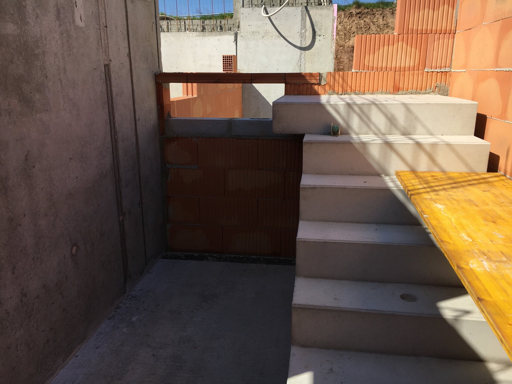 Construction site: concrete walls, white stairs, wooden board on the right, orange brick wall behind.