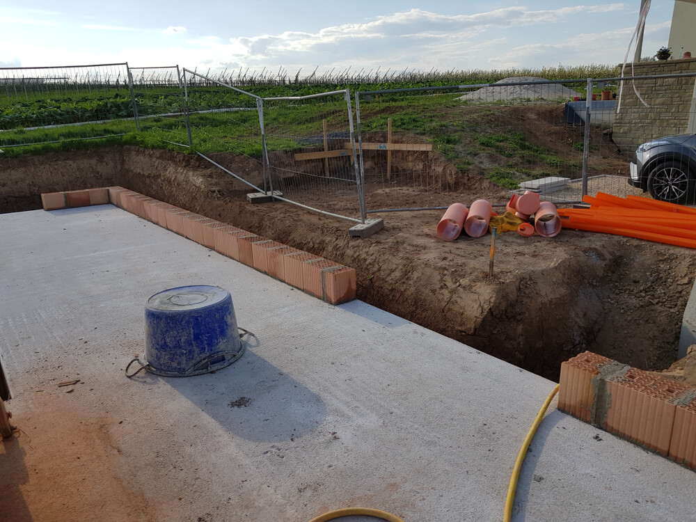 Construction site: concrete floor, brick edge, orange pipes, blue watering can, safety barrier in the background.