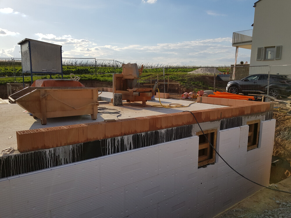 Construction site with concrete foundation, concrete mixer, construction equipment; house and car in the background.