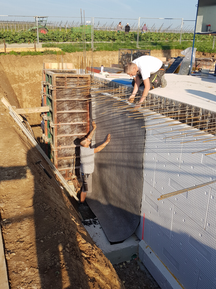 Two construction workers placing a gray insulation board against a foundation wall; reinforcement is laid on top.