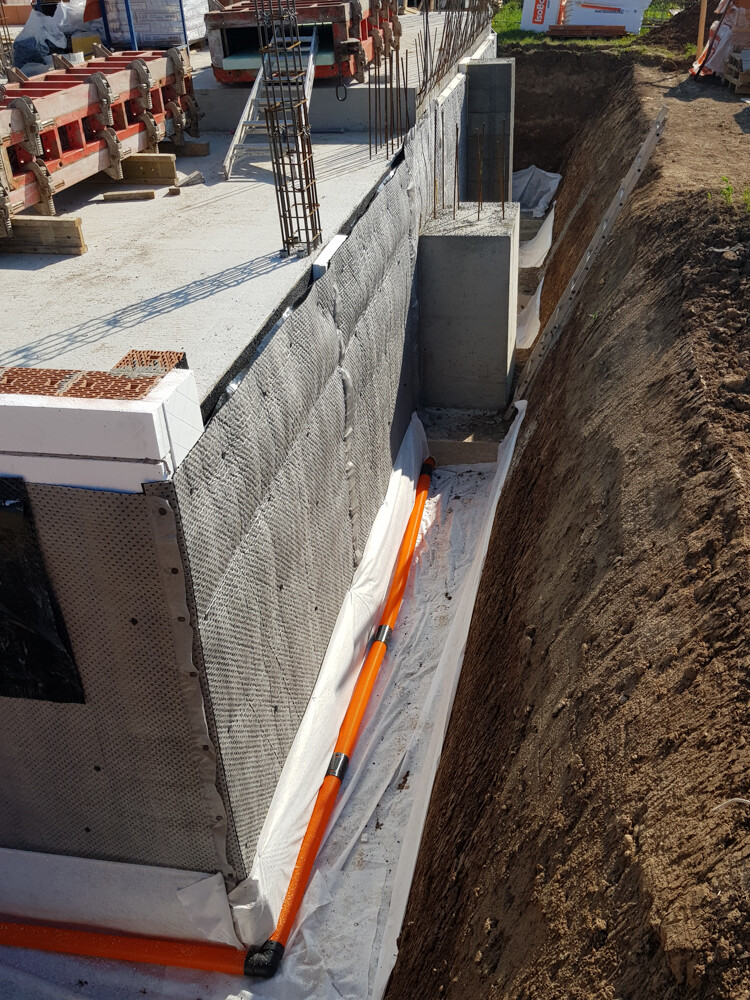 Foundation structure: concrete wall with reinforcement, orange pipe laid in the trench next to the wall.