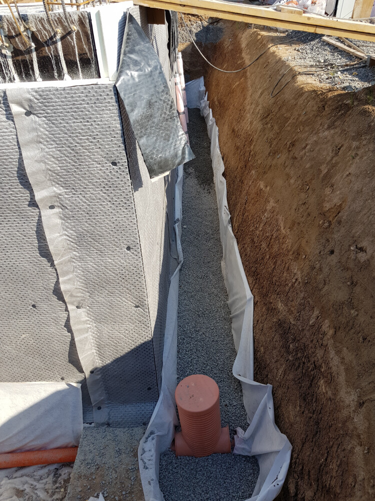 Excavation at the foundation: gravel base, waterproofing membrane on the wall, red pipe stub in the trench.