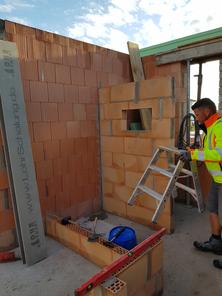 Construction site with orange brick wall, window opening, worker in high-visibility jacket next to ladder.