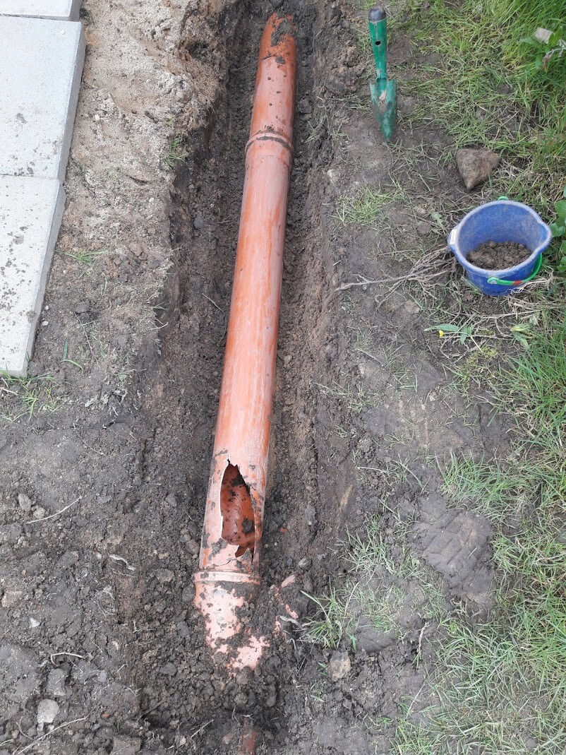 Orange pipe in trench, damaged with a hole; shovel and bucket next to it.