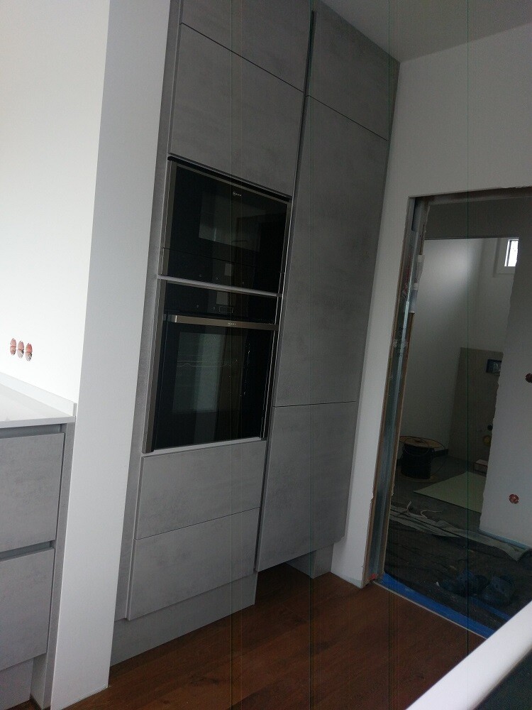 Modern kitchen with grey tall cabinets and built-in ovens as well as wood flooring.