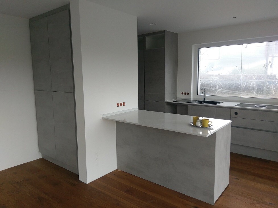 Modern kitchen: grey fronts, concrete-look island, wood floor, sink by the window.