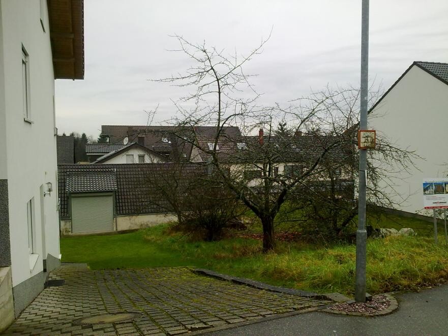 Exterior view of a residential area: white house on the left, paved forecourt, grass and trees.