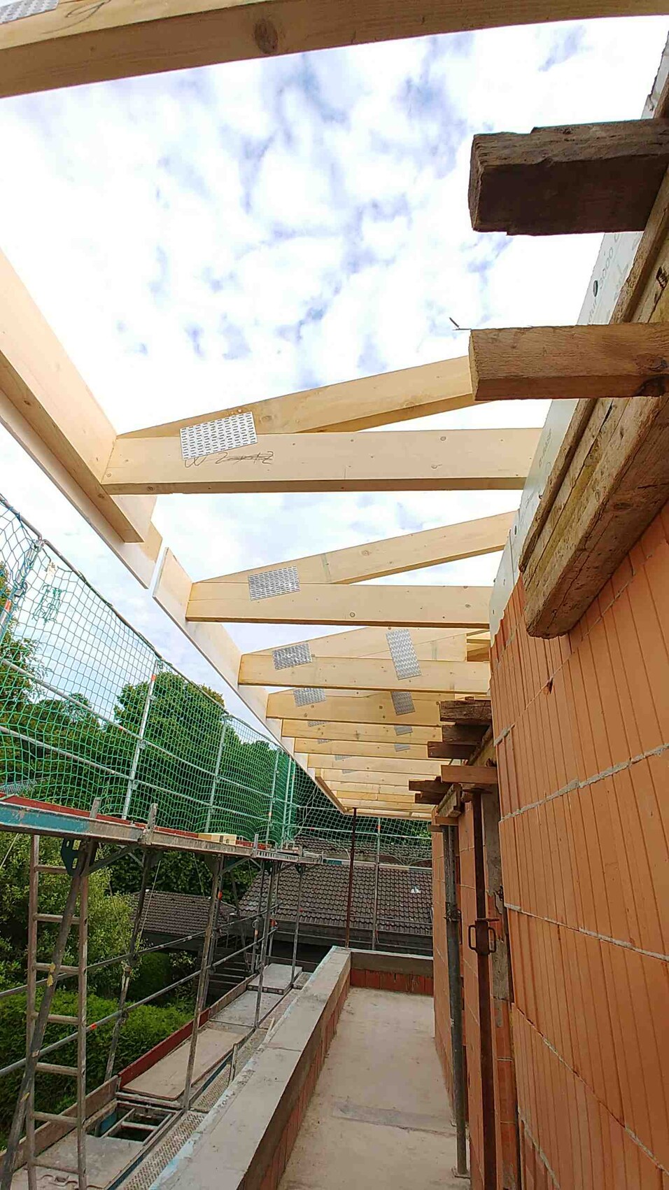 Baugerüst neben Backsteinwand; Holz-Dachträger werden montiert, offener Himmel.