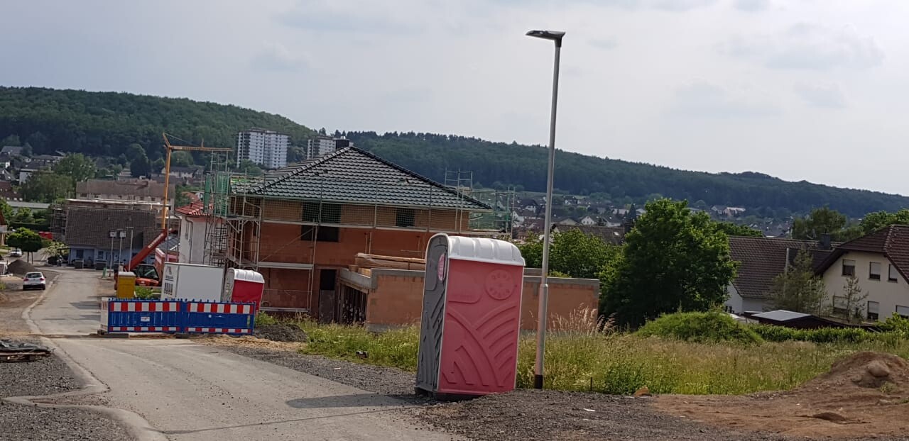 Construction site: pink portable toilet cabin in front of scaffolding, barrier tape, houses in the background.
