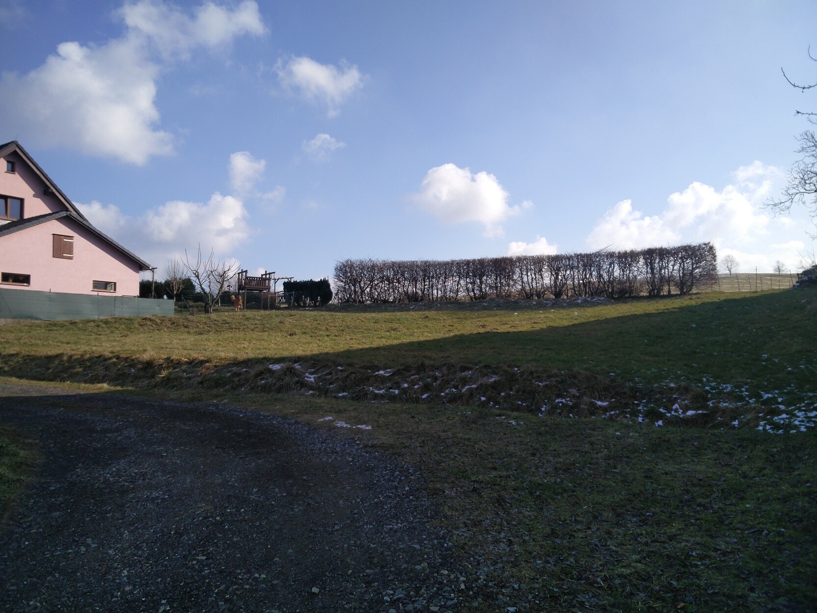 Außenansicht eines pinken Hauses mit Garten, Hecke und offener Landschaft unter blauem Himmel.
