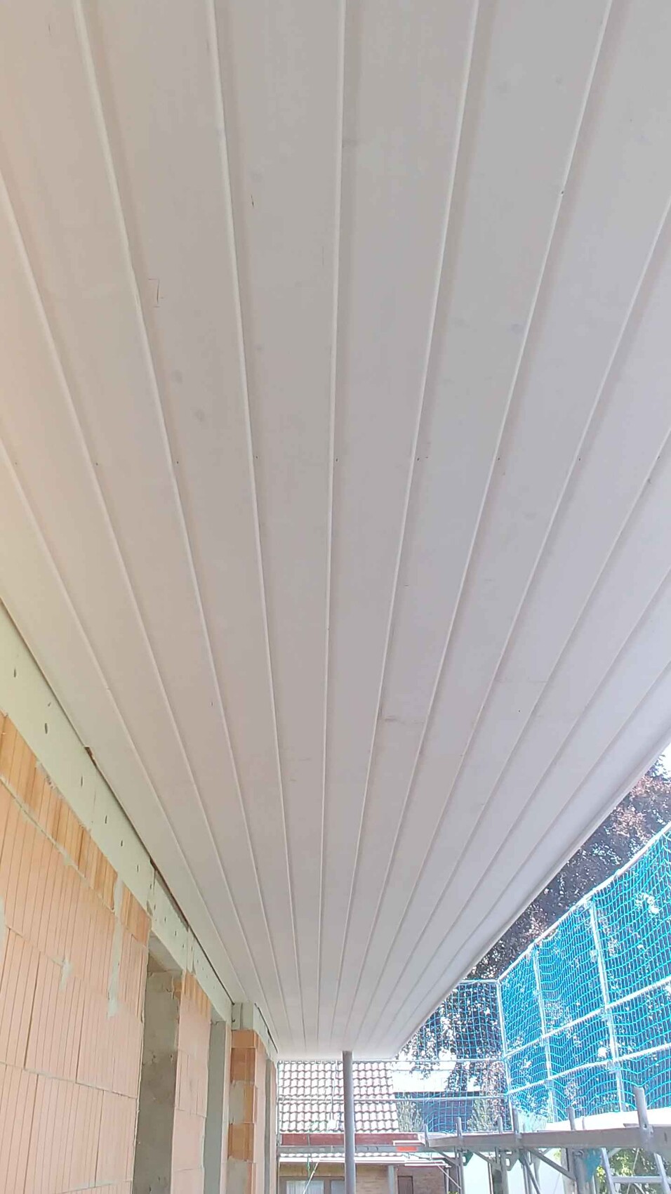 Weiße Dachverkleidung aus Paneelen einer Veranda, Baugerüst und Bruchsteinwand sichtbar