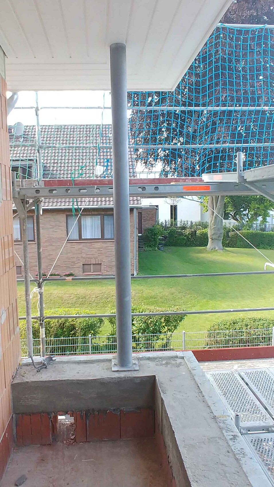 Außenbereich einer Baustelle mit Gerüst, blauem Schutznetz und angrenzendem Garten