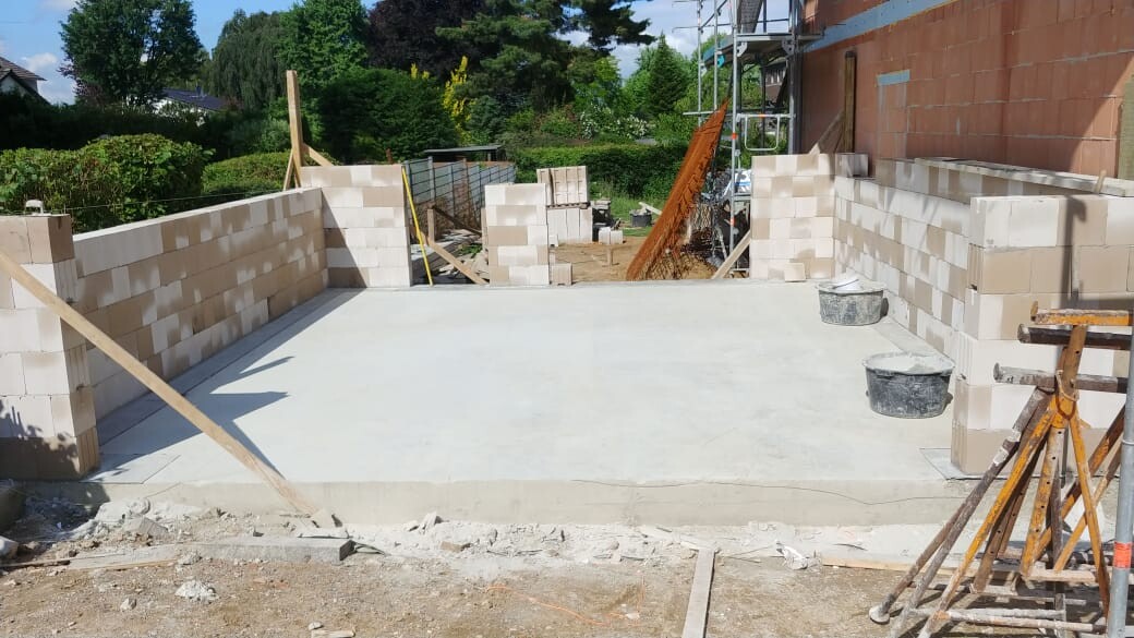 Baustelle: ebene Betonbodenplatte, teilweise gemauerte Blockwände, Baugerüst und Eimer.