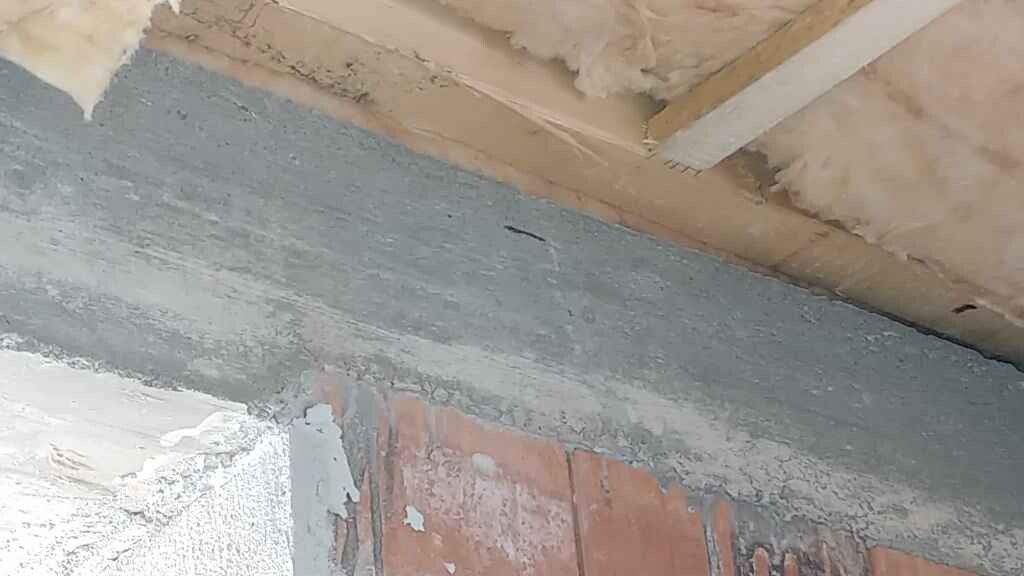Interior view of an unfinished wall: gray plaster, bricks underneath, wooden beam ceiling with insulation.