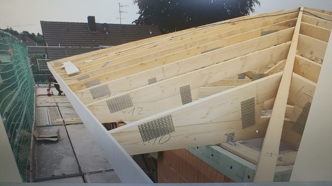 Section of wooden roof truss with metal plates on load-bearing beams; scaffolding on the left.