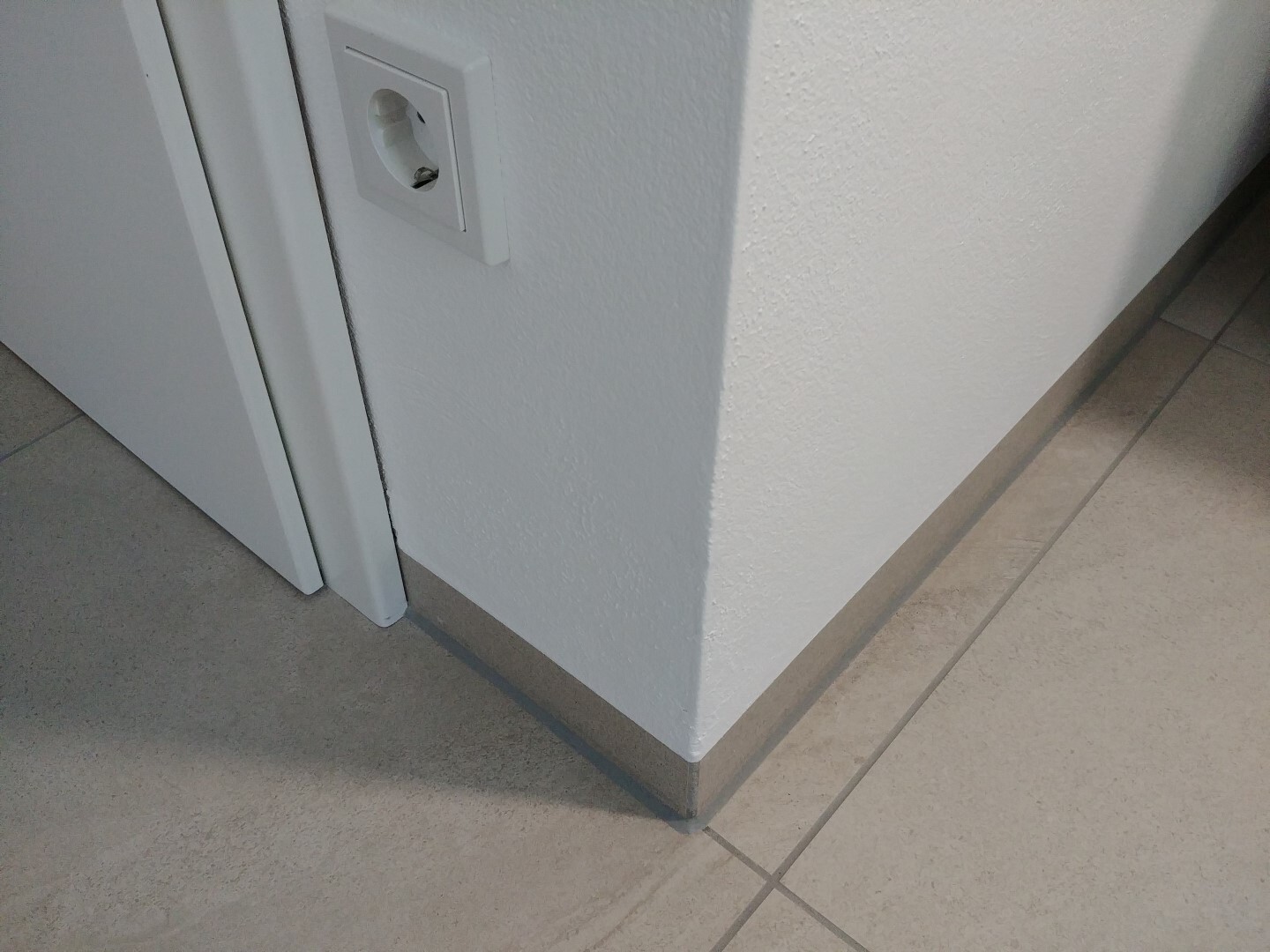 Weiße Innenwand-Ecke mit Türrahmen, Bodenfliesen und Wandsteckdose.